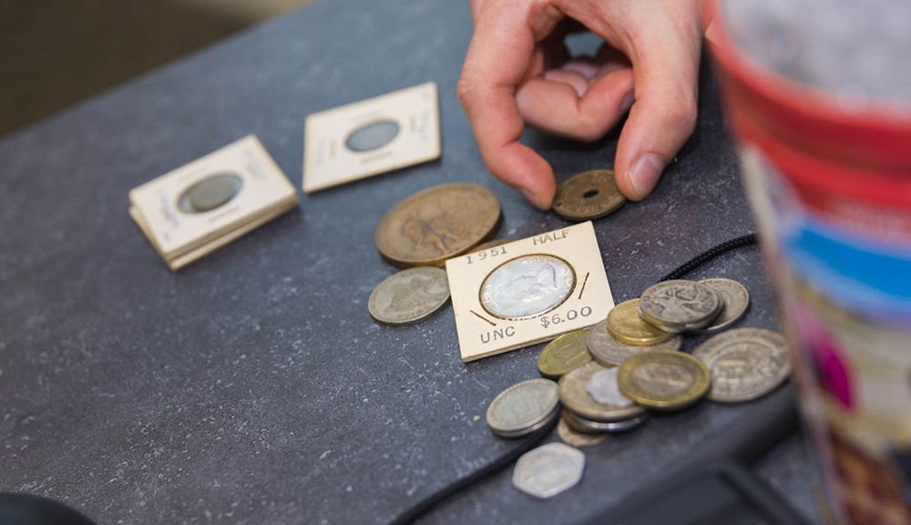 A hand reaches from out of the frame to pick up one of several old coins on a tabletop.