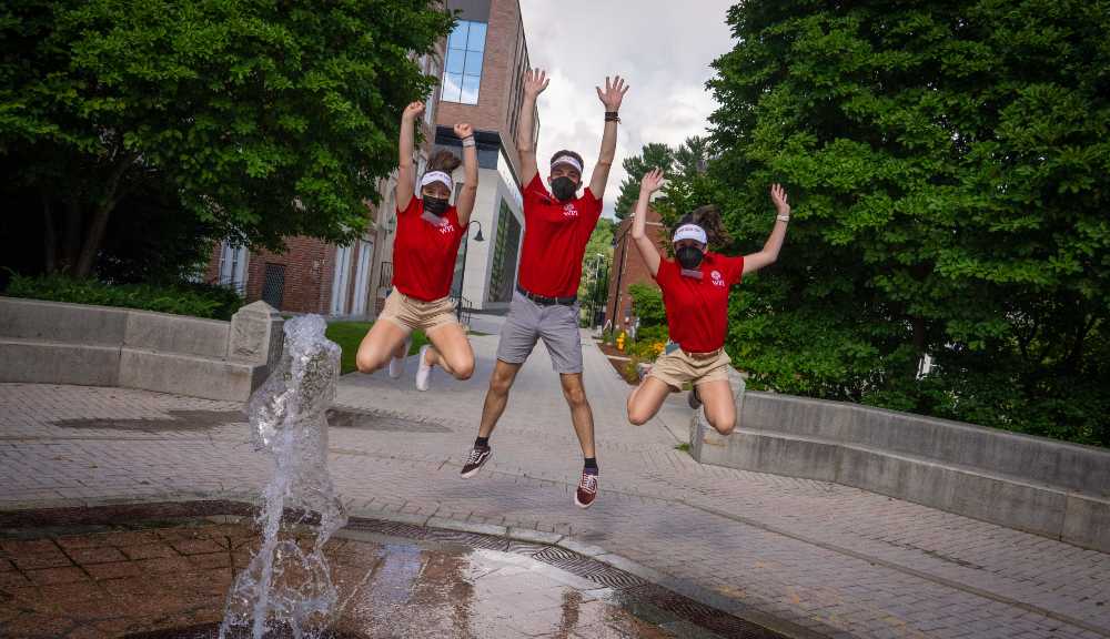Three orientation leaders jump in unison behind the fountain.