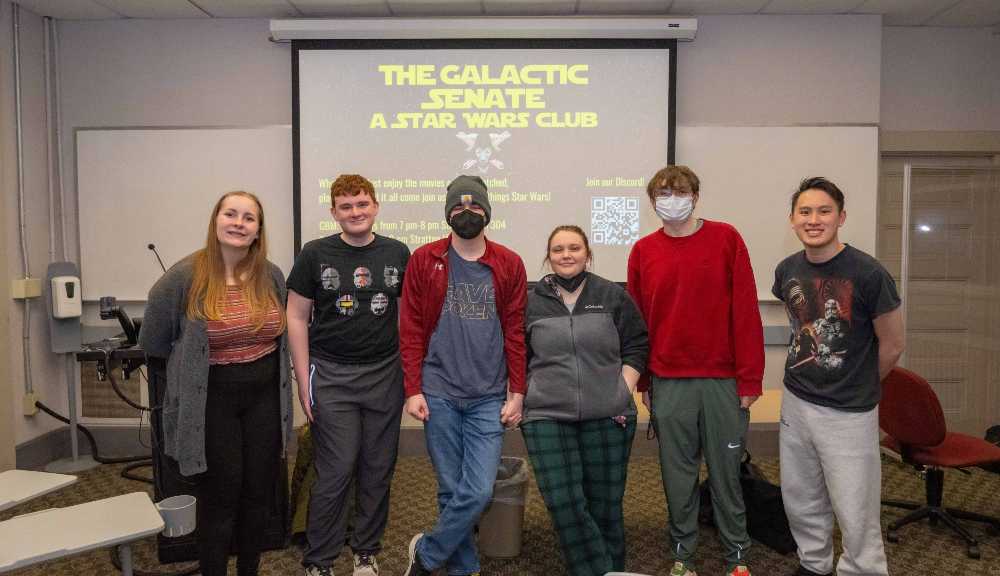 Members of the Star Wars Club smile for a photo during a trivia night event. The background projector reads "The galactic senate, a star wars club"