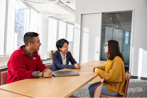 Two students, one in red top and one in yellow top, sit talking with President Wang at a conference table