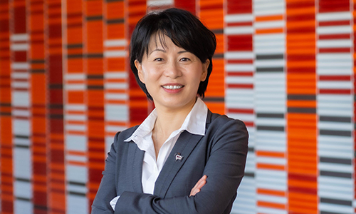head shot of Grace Wang in a gray blazer standing in front of a wall with red and gray subway tiles