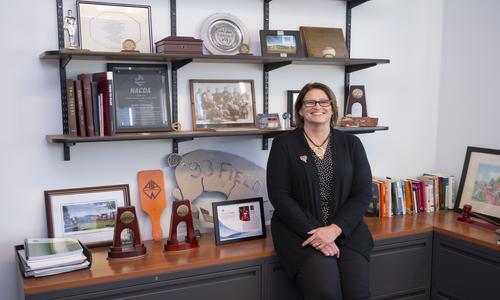 Dana Harmon sitting at the edge of a low desk in front of many awards on the wall and on the desk
