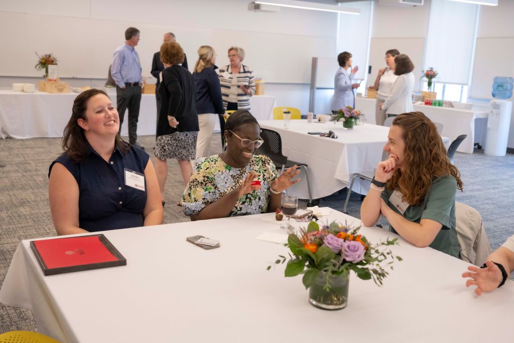 Women chatting at a table with people standing talking in the background at the Board of Trustees Outstanding Staff Awards luncheon