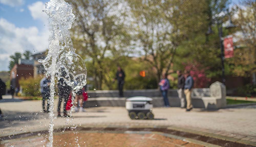 A photo of the fountain with students and a food delivery robot in the background.