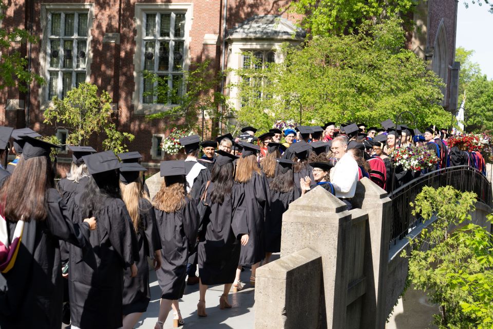 the backs of graduates in black caps and gowns proceeding across Earle Bridge for commencement