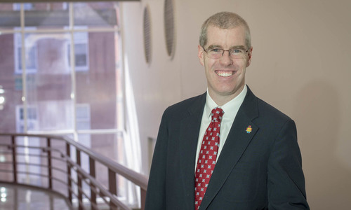 Neil Heffernan standing in front of a poster in a dark suit jacket and red tie