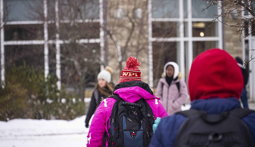 A student walks across campus through the snow with a WPI winter hat on.