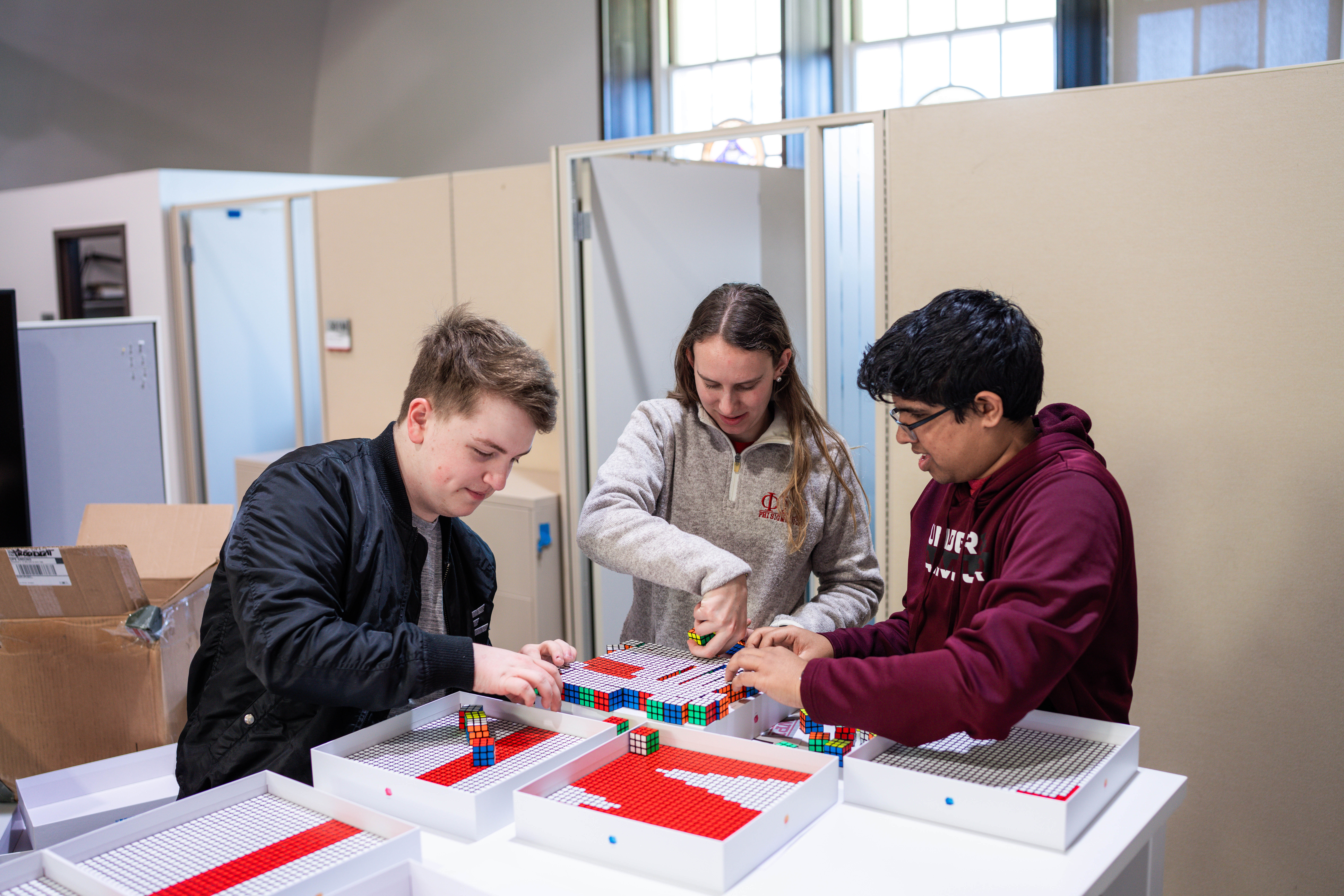 Three students arrange a Rubik’s Cube mosaic at a table.
