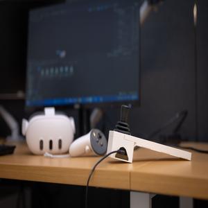 A virtual reality headset and two joystick style controllers placed on an office table, in front of a blank computer screen.