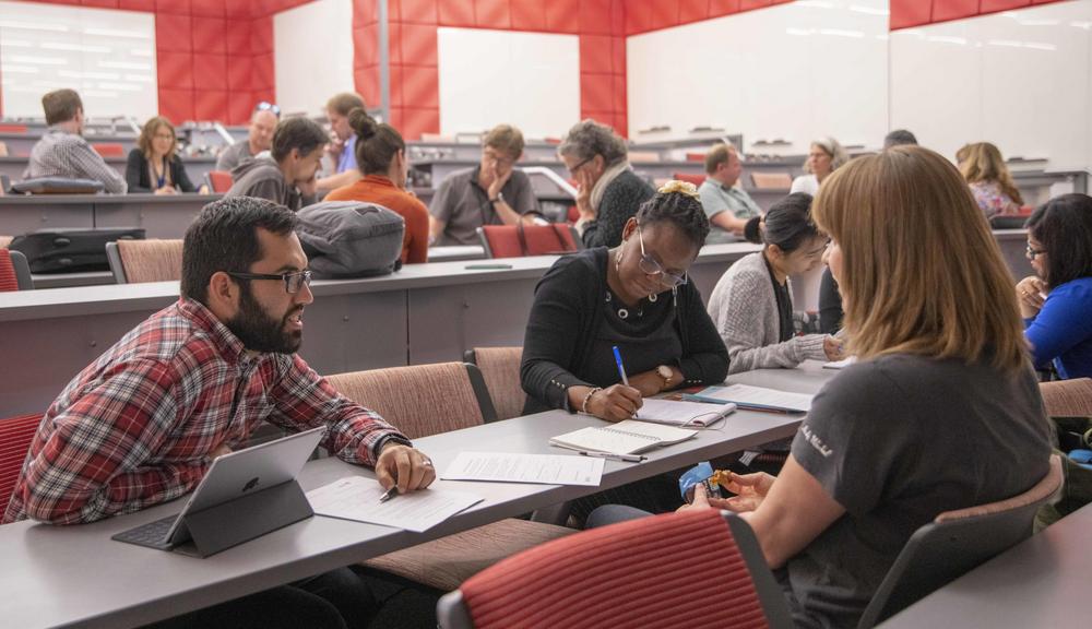 bearded man leans forward talking to a woman while people in background take notes during a session at the Institute on Project-Based Learning