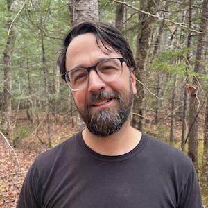 WPI Professor of Philosophy and Global Studies Geoffrey Pfeifer stands in a forest, smiling