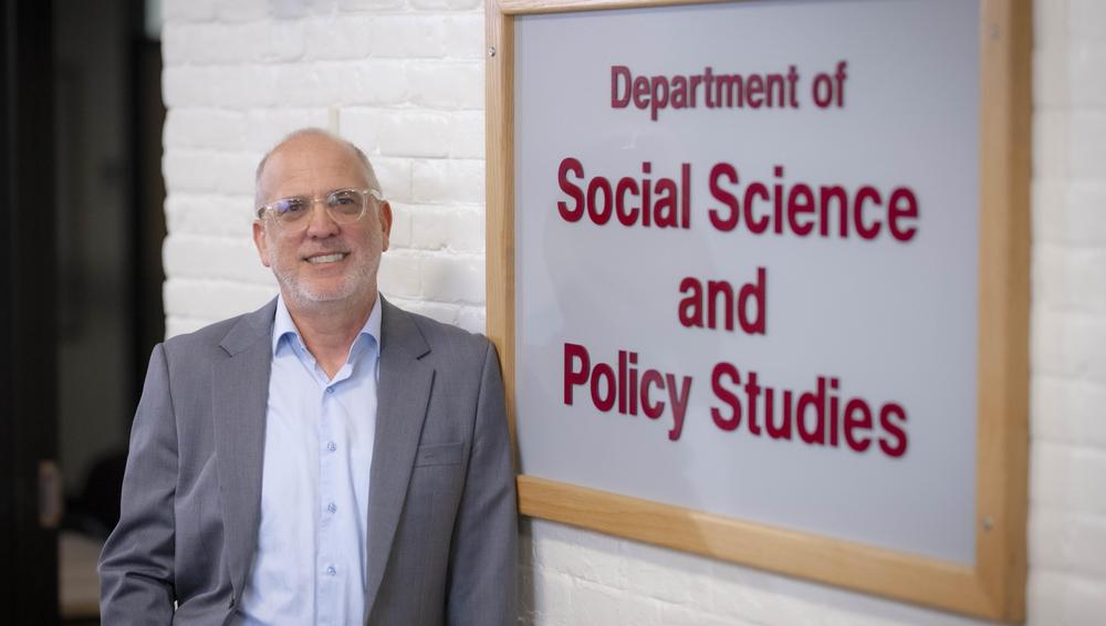 WPI researcher Robert Krueger leans against a sign for the Department of Social Science and Policy Studies. He is using AI to identify economic development opportunities.