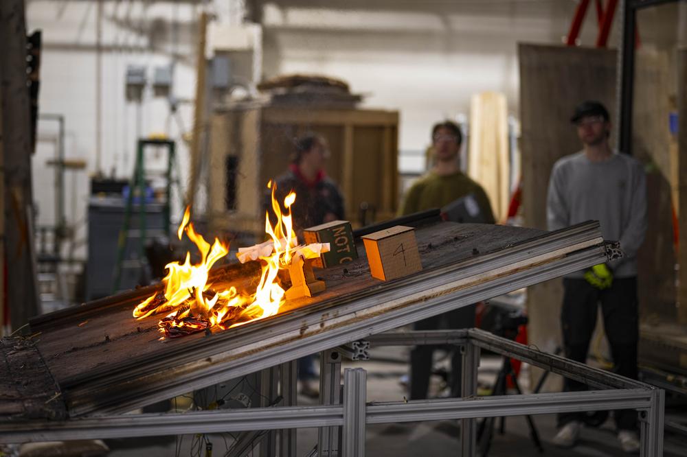 Cardboard models of houses and trees burn on a burn table inside a fire protection engineering laboratory at WPI as 3 students look on in the background to observe the experiment