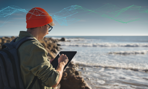 Person standing on a beach studies the ocean while using a tablet device.